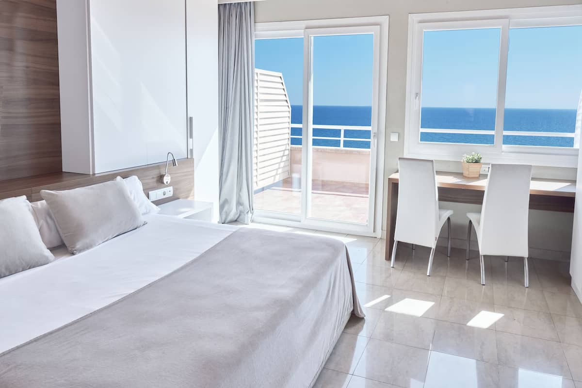 A modern hotel room with a large bed, a desk with chairs, and a view of the ocean through large windows.