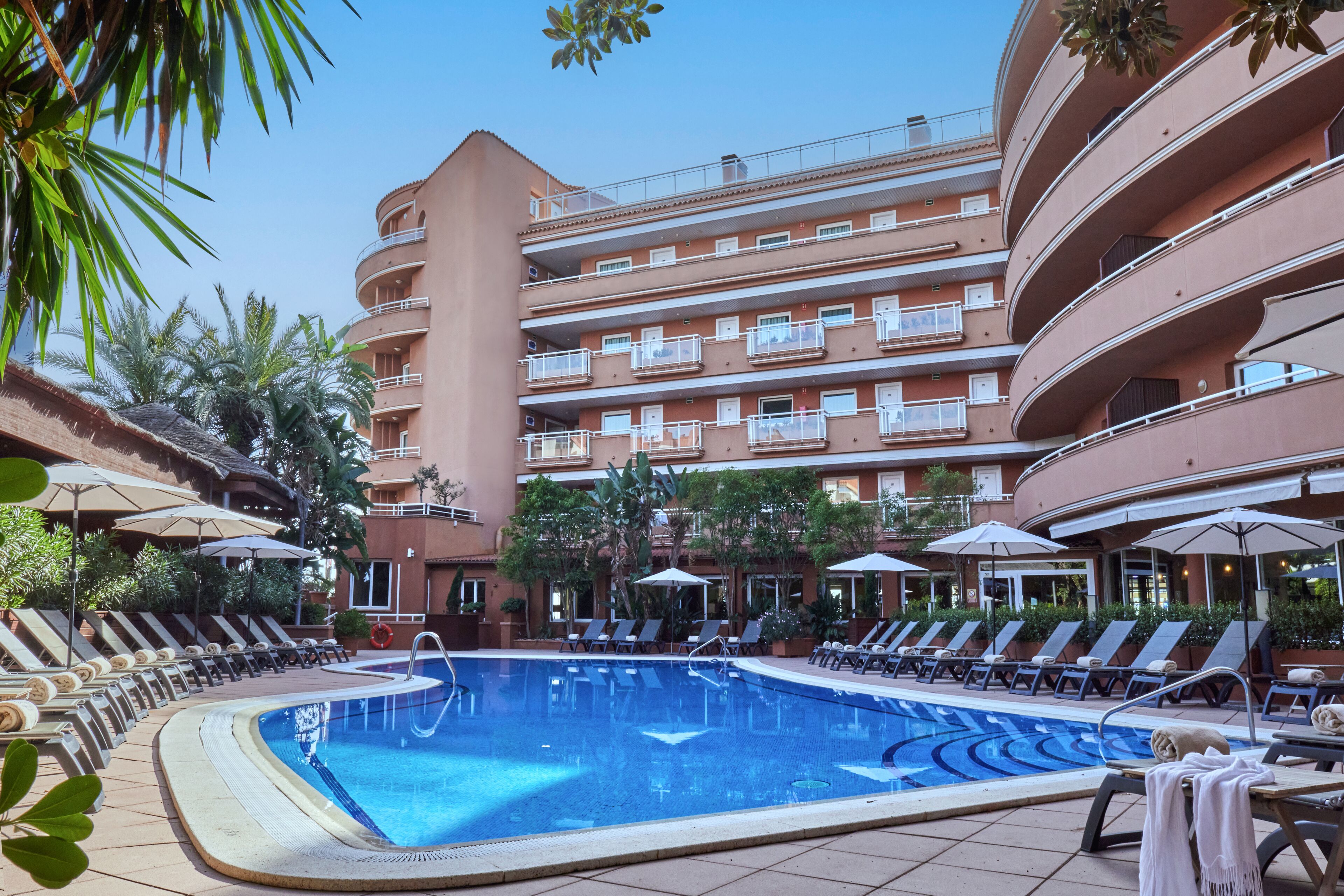 A hotel pool area with multiple lounge chairs and umbrellas, surrounded by a multi-story building with balconies.