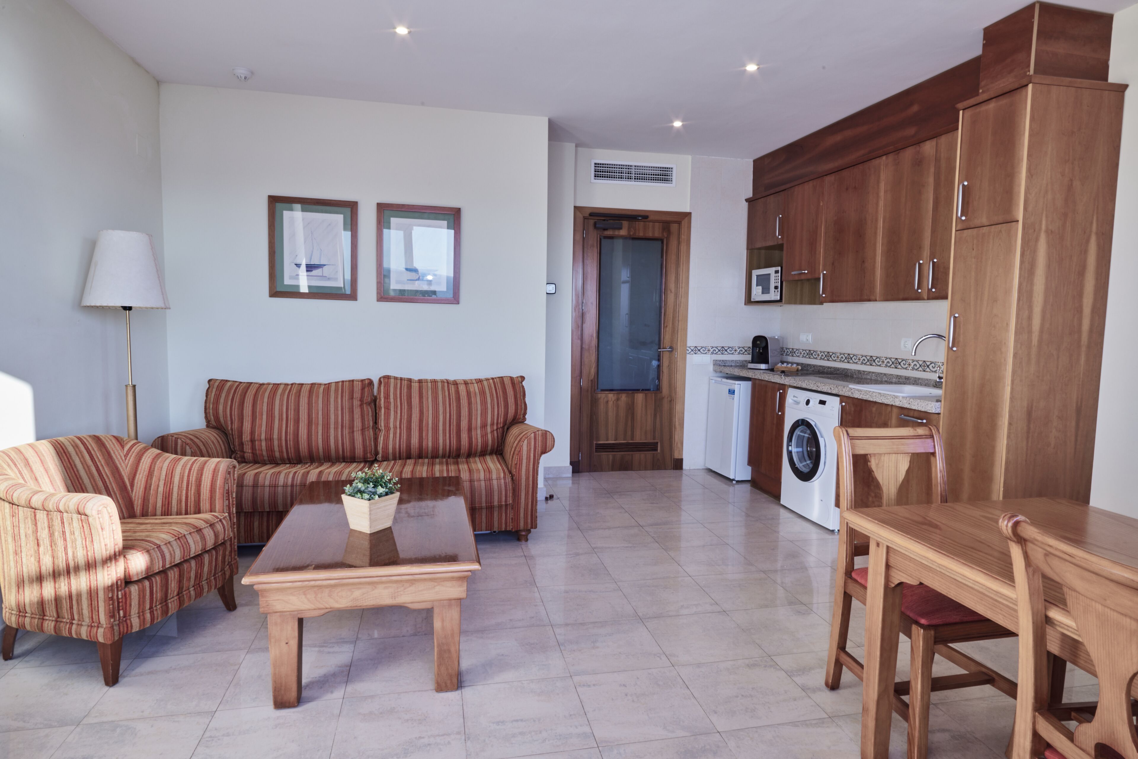 A living room with a sofa, a coffee table, and a dining table. There is a washing machine and a microwave in the kitchen area.