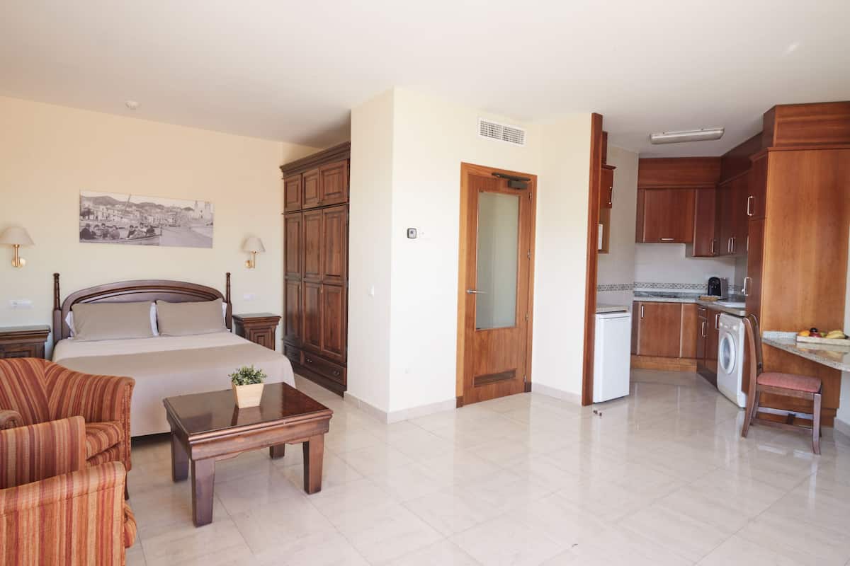 A hotel room with a bed, a sitting area with a chair and table, a kitchen area with a washing machine, and a dining area with a table and chairs.