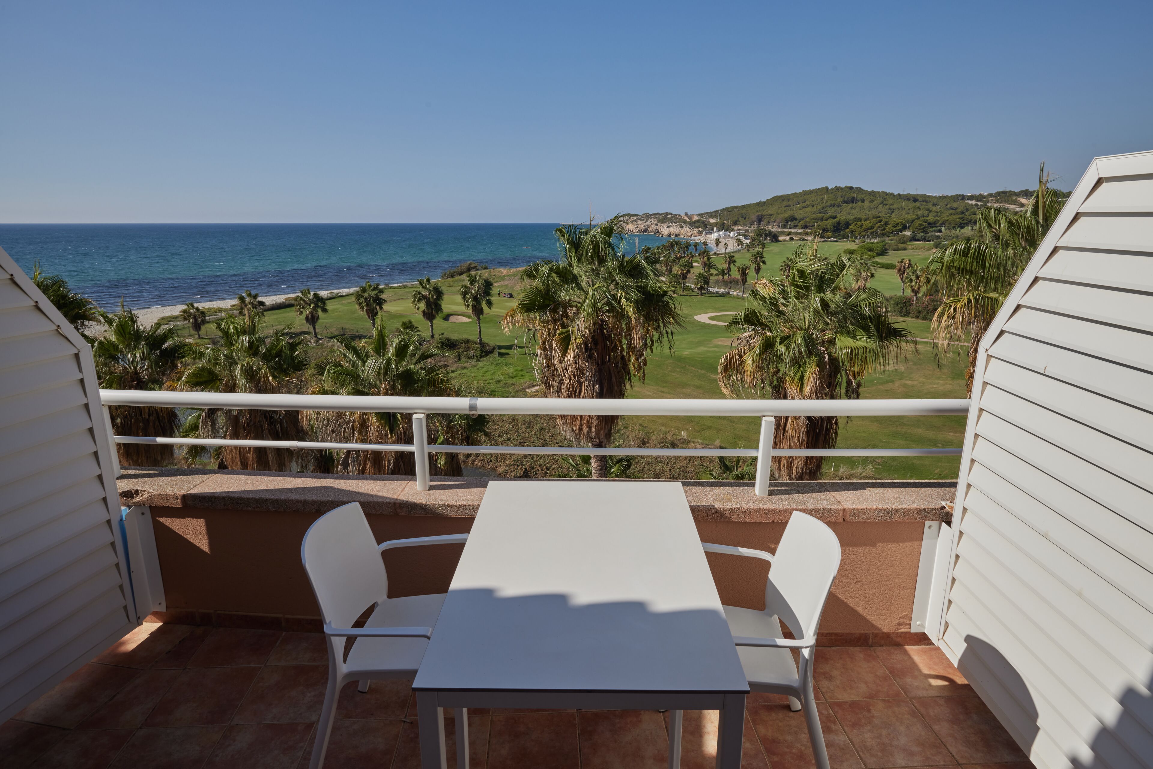 A balcony with a white table and chairs, overlooking a golf course and the ocean.