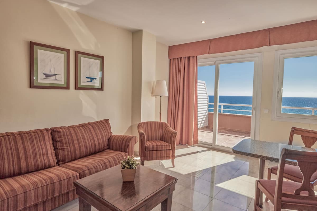 A hotel room with a sofa, armchair, table, and a balcony with a view of the sea.