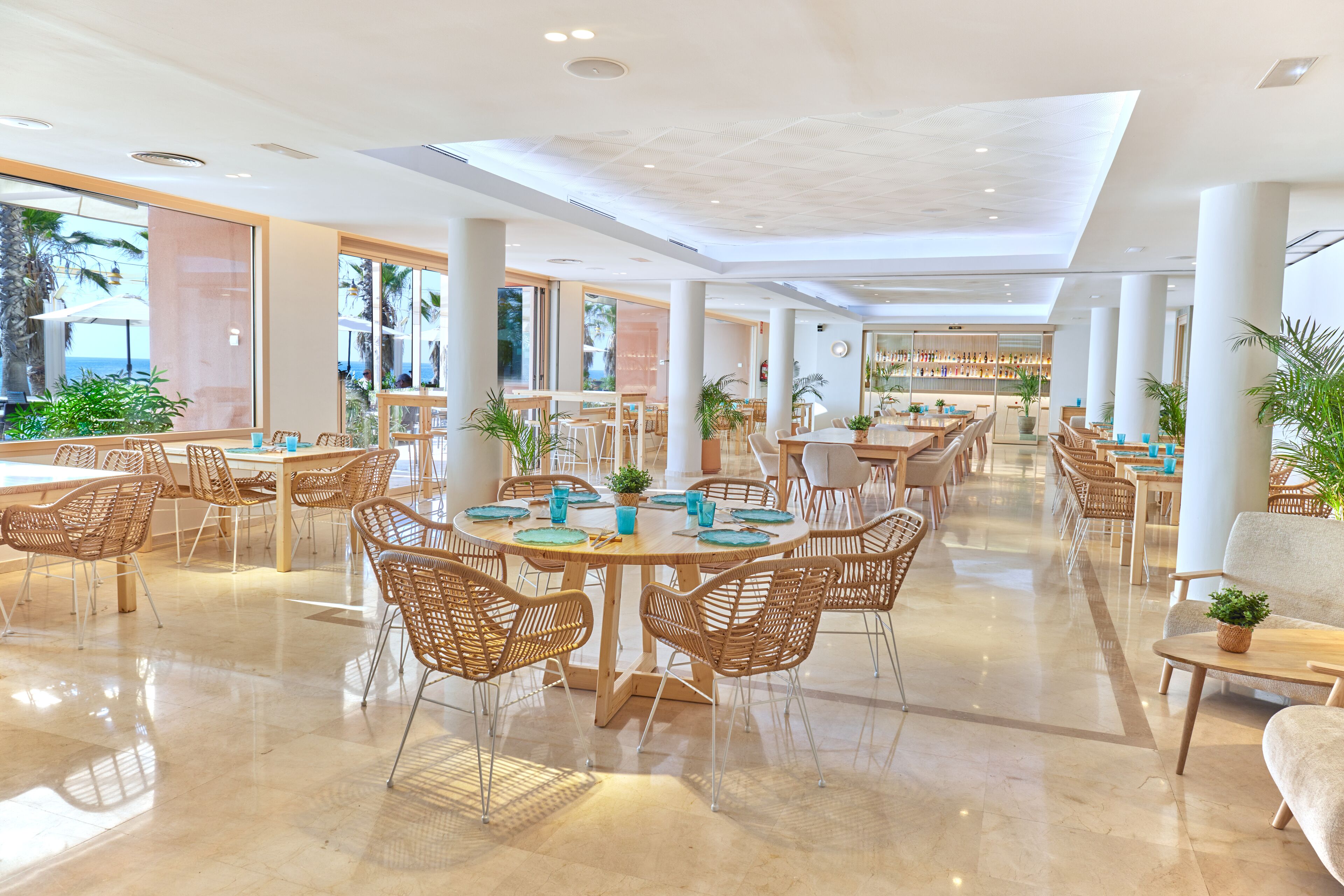 A spacious dining area with round tables, wicker chairs, and potted plants.