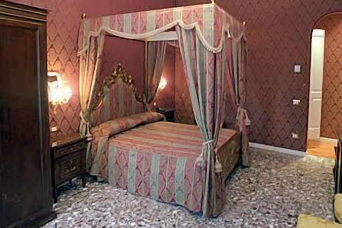 A four-poster canopy bed with a patterned bedspread, a nightstand, and a wall-mounted lamp.