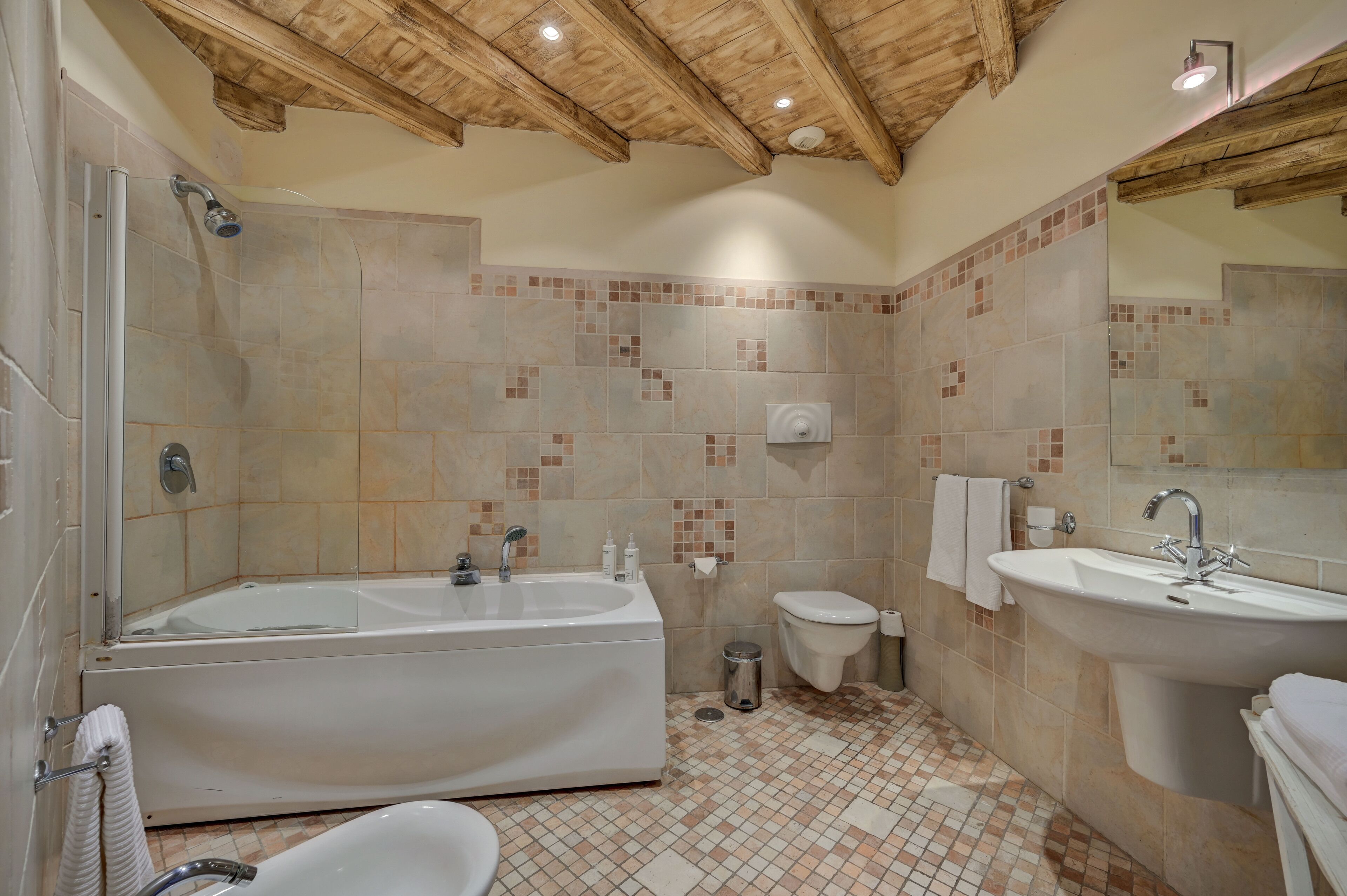 A bathroom with a bathtub, toilet, and sink, featuring tiled walls and wooden ceiling beams.