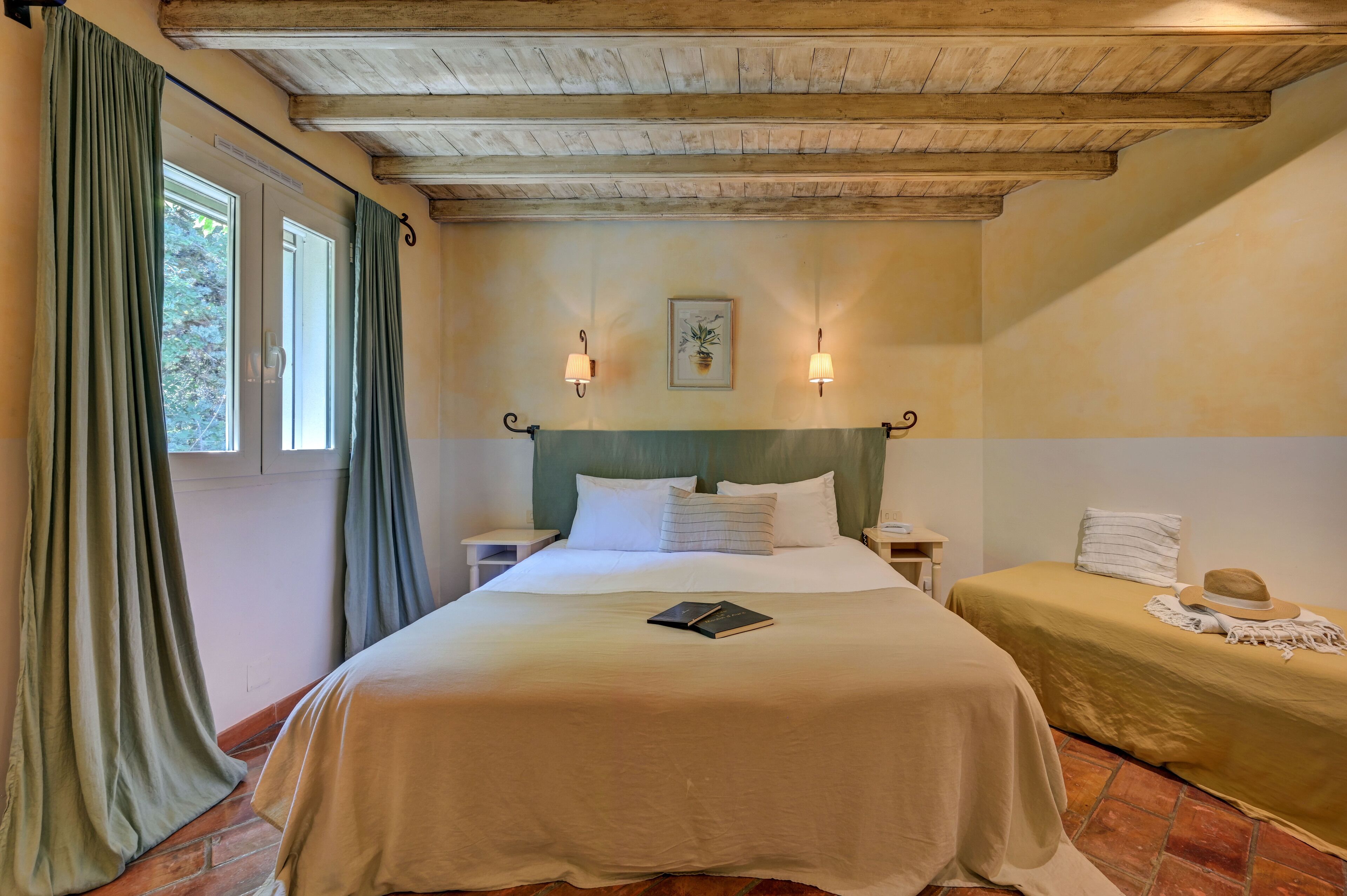 A double bed with a book, two bedside tables, a headboard, a window with curtains, and a wall-mounted lamp.