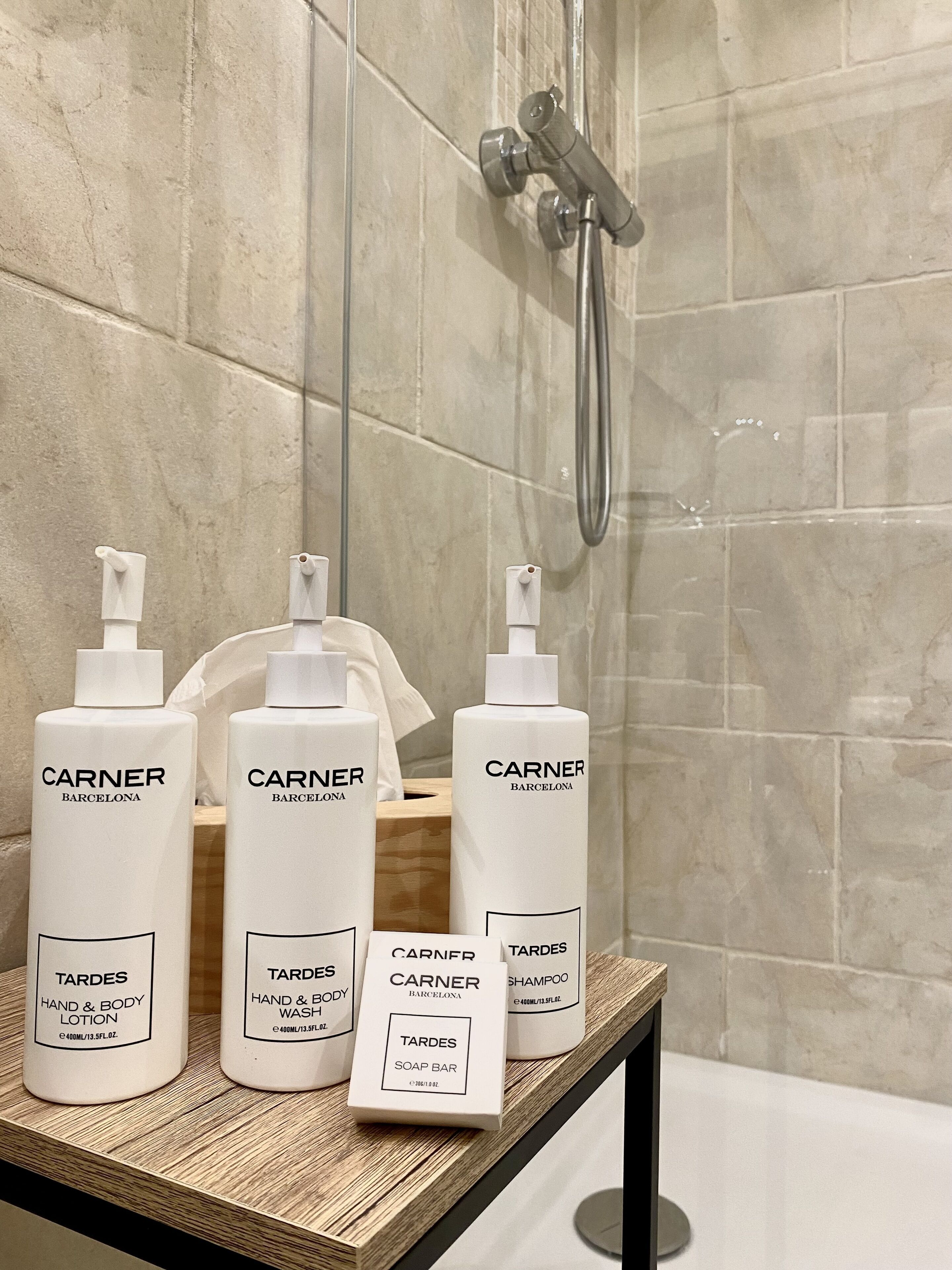A bathroom with a shower, a wooden shelf with three bottles of Carner Barcelona hand and body care products, and a soap bar.