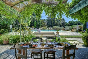 Un área de comedor al aire libre con una mesa lista para dos personas, rodeada de exuberante follaje y una piscina al fondo.
