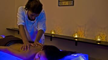 Eine Massageanwendung in einem schwach beleuchteten Raum mit einer Massageliege, einem liegenden Klienten, der mit dem Gesicht nach unten liegt, und einem Therapeuten, der eine Rückenmassage durchführt.