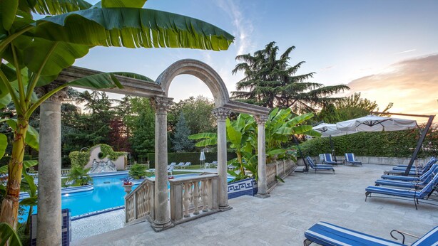 Zona de piscina con sillones reclinables y sombrillas, rodeada de vegetación y columnas arquitectónicas.