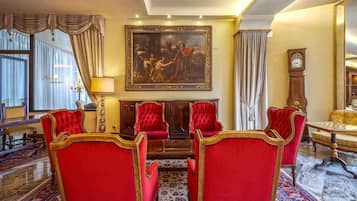 Uma sala de jantar formal com cadeiras estofadas em vermelho, um quadro grande sobre a lareira e mobiliário clássico.