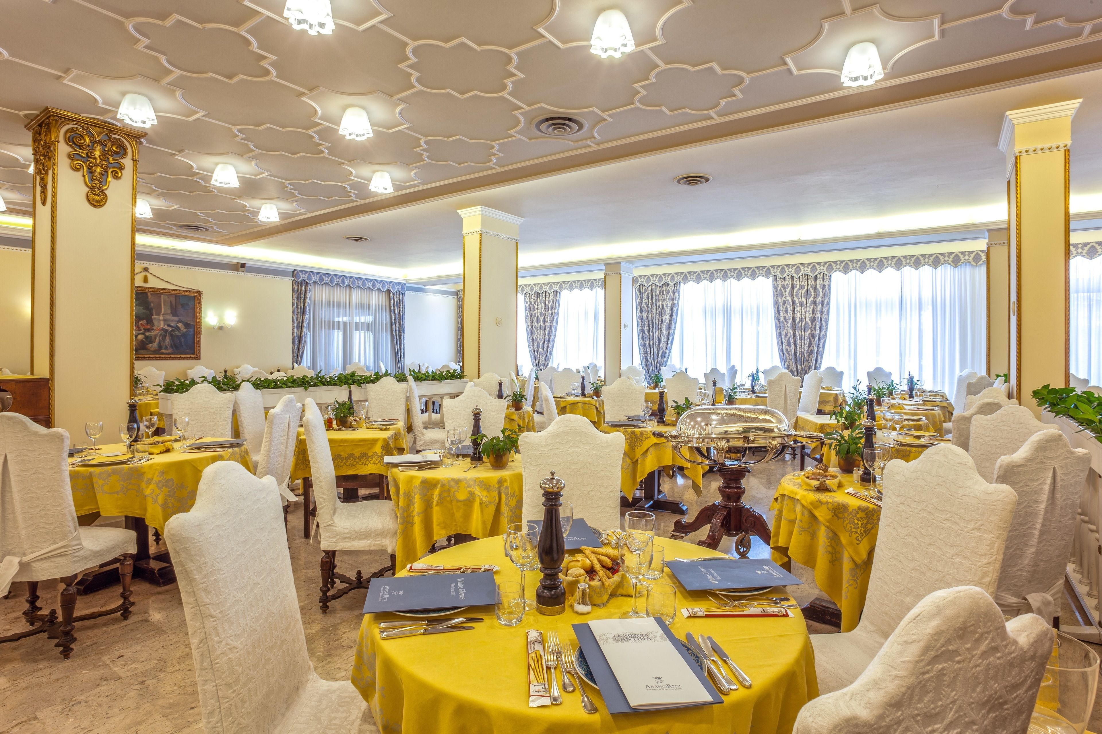 A formal dining room with multiple tables set for service, featuring white chairs and yellow tablecloths.