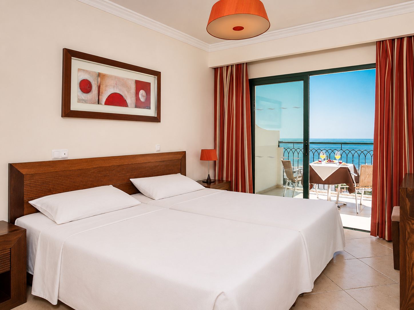 A hotel room with a double bed, a framed abstract painting, a balcony with a table and chairs, and a view of the sea.