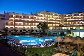 A hotel with a large swimming pool surrounded by lounge chairs and umbrellas, multiple balconies, and a well-lit facade.