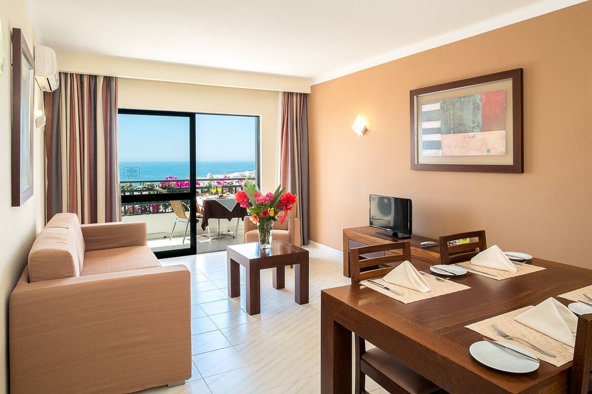 A hotel room with a sofa, dining table set for two, a TV, and a balcony with a view of the sea.