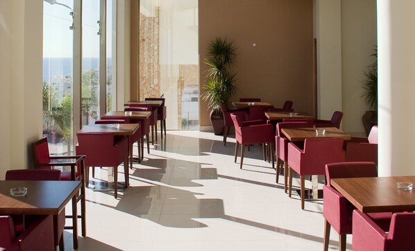 A spacious indoor area with multiple tables and chairs, large windows offering a view of the city, and a stone wall.