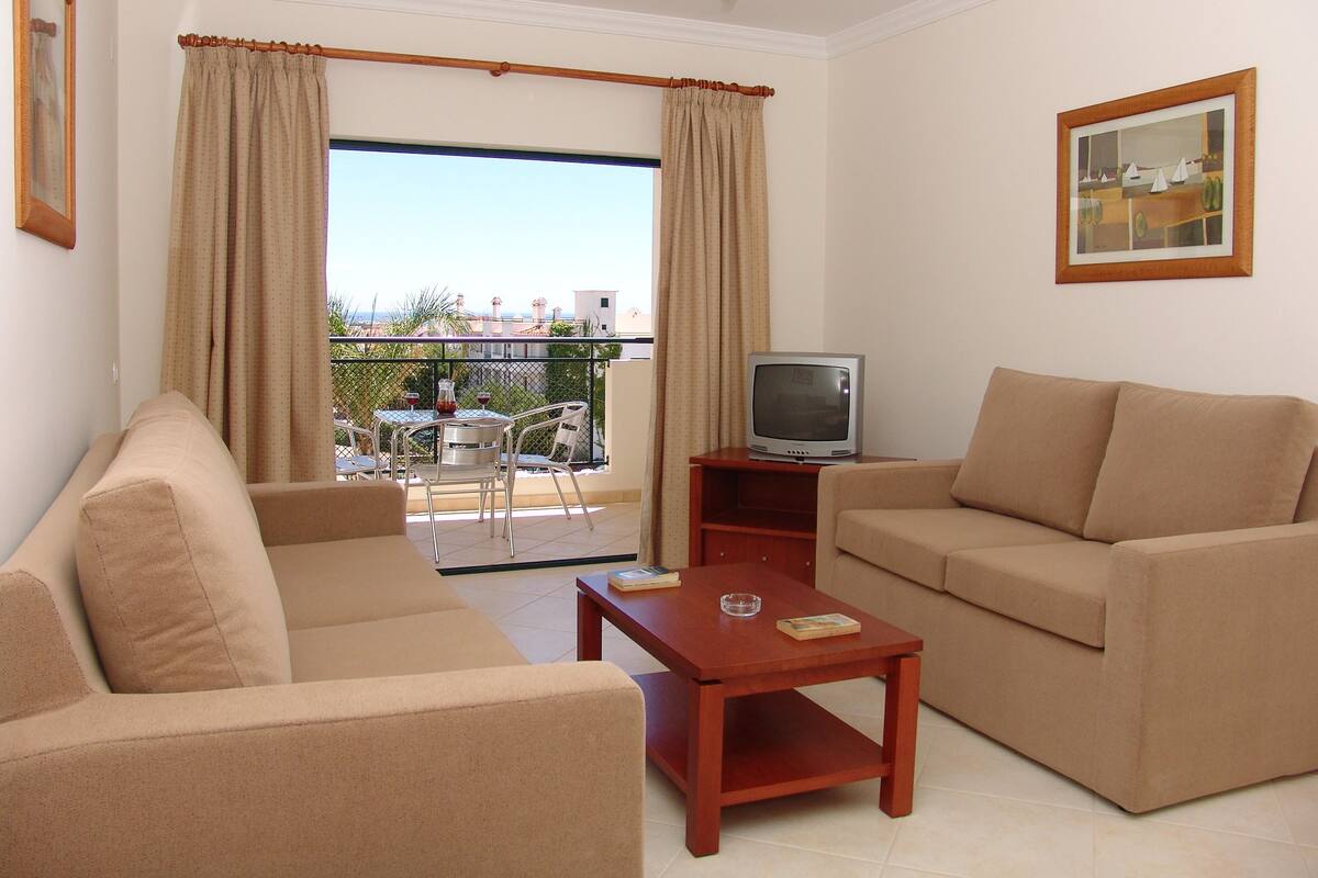 A living room with a sofa, a TV on a stand, a small table, and a balcony with a view.