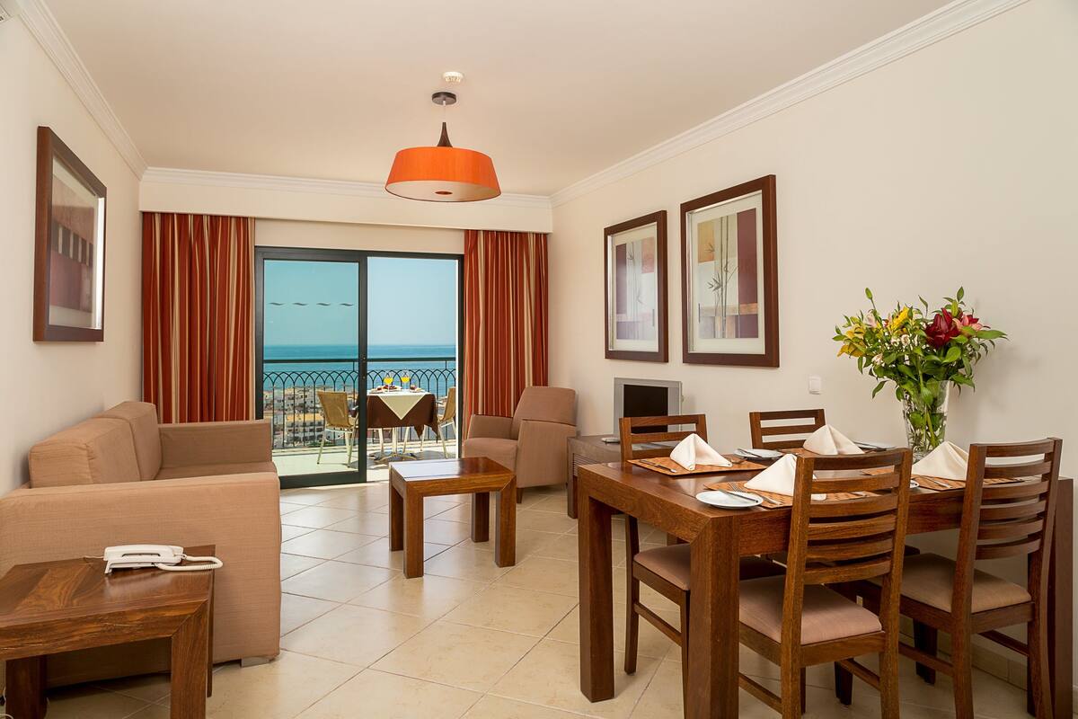 A hotel room with a dining area, a sofa, a wooden table, and a view of the sea through the balcony doors.