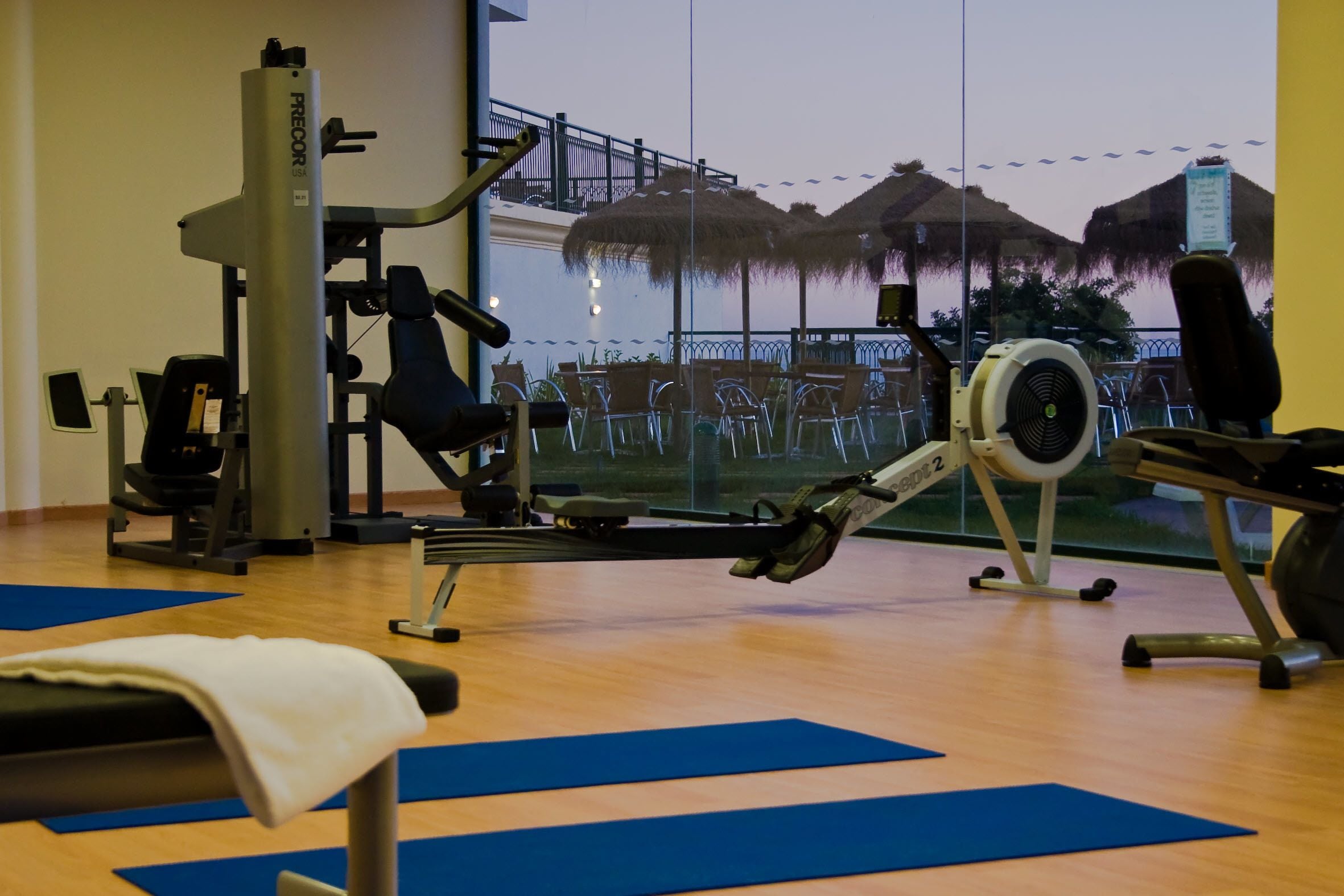 Un gimnasio con equipo de ejercicio, colchonetas azules y vistas a una playa con sombrillas.