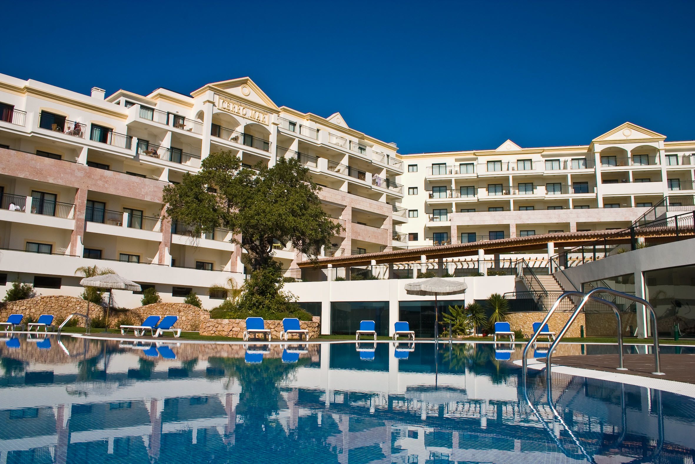 A hotel with a swimming pool, blue lounge chairs, and a large tree in the foreground.