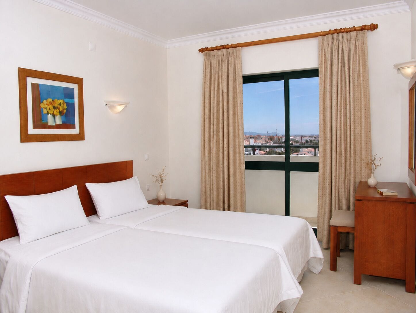 A hotel room with a bed, a desk, a chair, a window with curtains, a vase with flowers, and a framed picture on the wall.