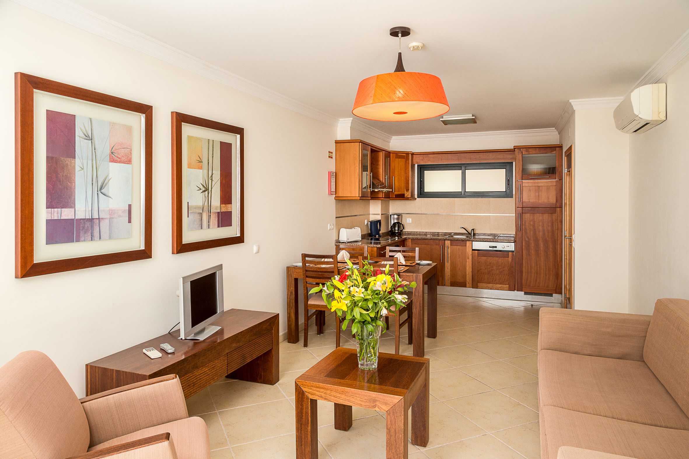 A modern living room with a TV, wooden furniture, a dining table with chairs, and a kitchen in the background.