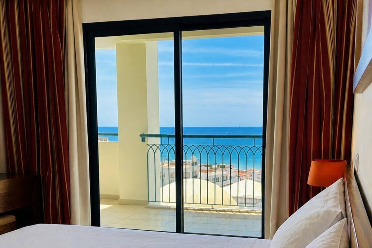 A hotel room with a bed, curtains, a view of the beach, and a balcony.