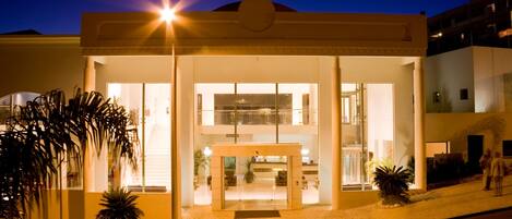 A hotel entrance with a glass facade, a reception desk, and a staircase leading to the upper floors.