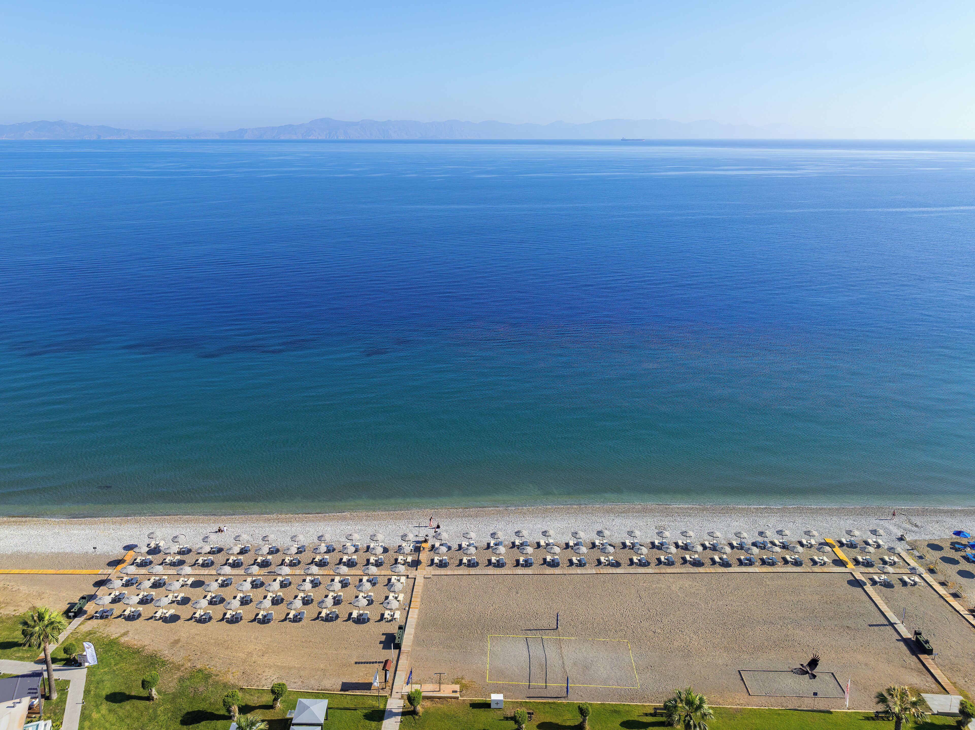 Vista aérea de una playa con filas de tumbonas y sombrillas, una cancha de voleibol y una vista despejada del mar y montañas distantes.