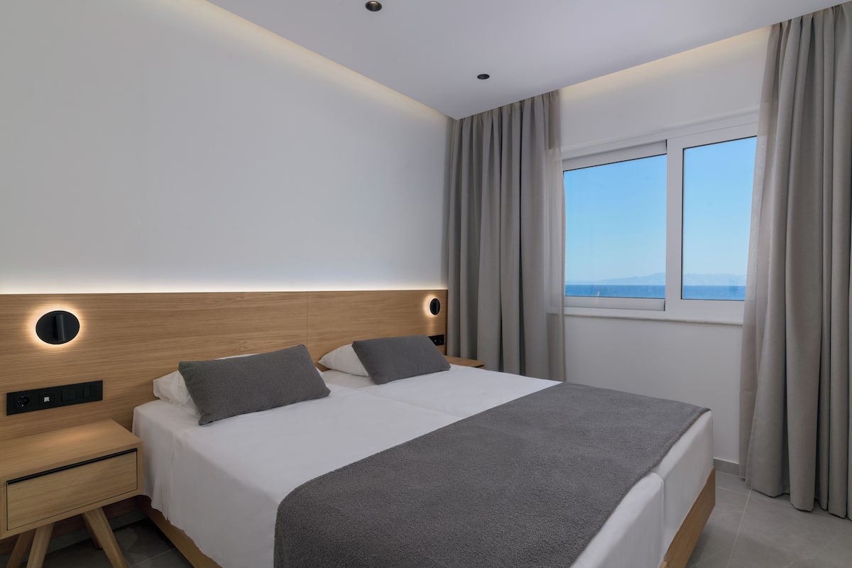 A modern hotel room with a large bed, a bedside table, a built-in nightstand, and a window with curtains.