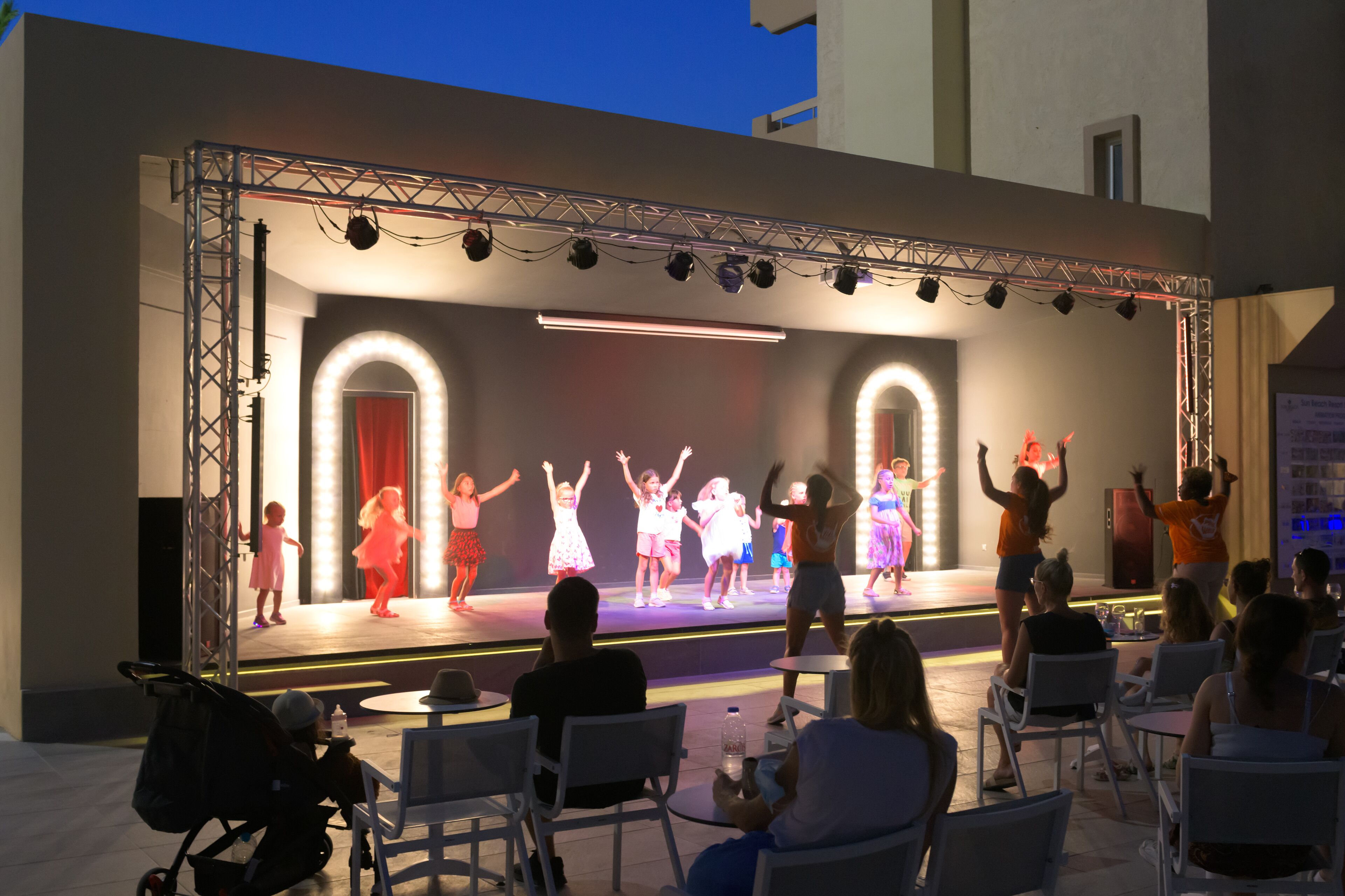 An outdoor stage with a performance, surrounded by seated chairs and tables.