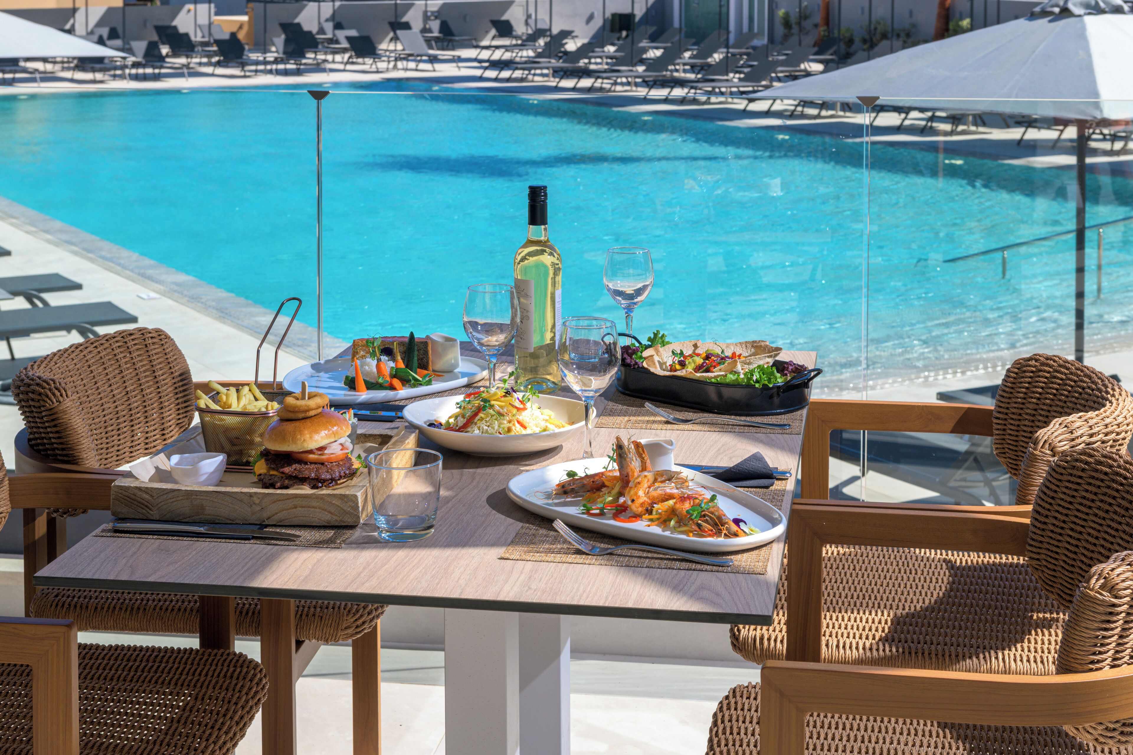 A dining table with plates of food, a bottle, and glasses, overlooking a pool with lounge chairs.