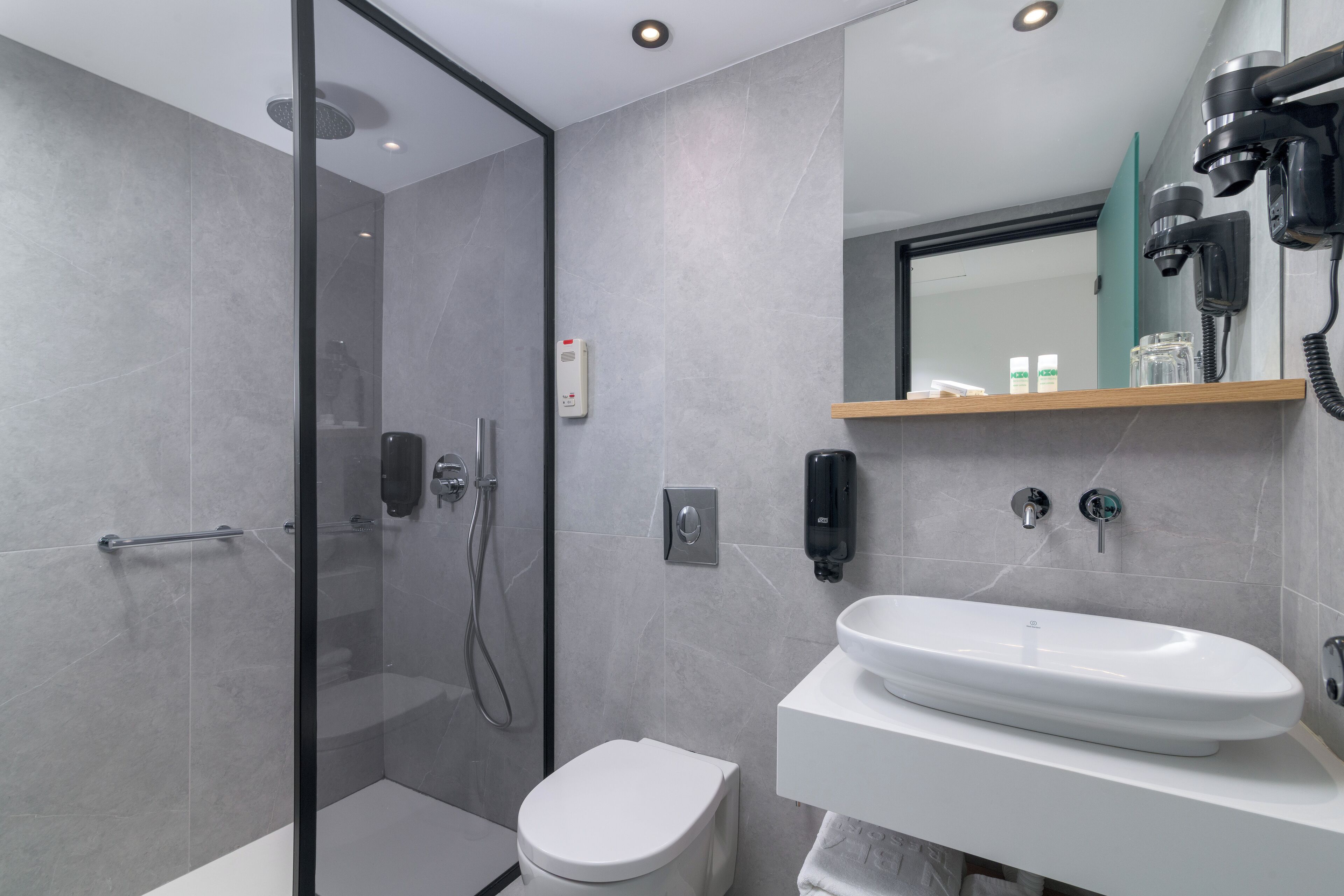 A modern bathroom with a glass shower enclosure, a white toilet, a sink with a mirror above, and a hairdryer mounted on the wall.