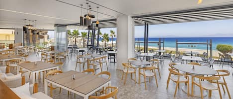 A modern restaurant with outdoor seating overlooking a beach, featuring wooden tables and chairs, large windows, and a view of the ocean.