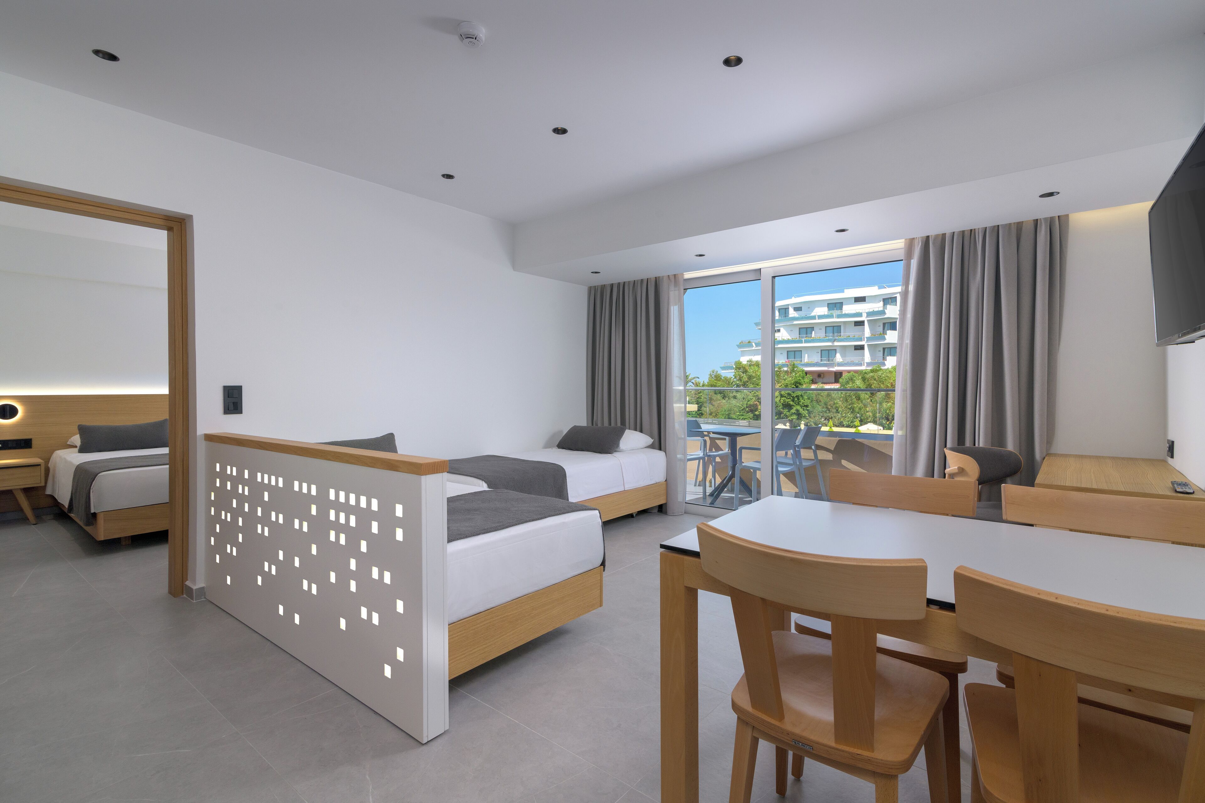 A modern hotel room with two beds, a dining area, and a balcony with a view of buildings and trees.