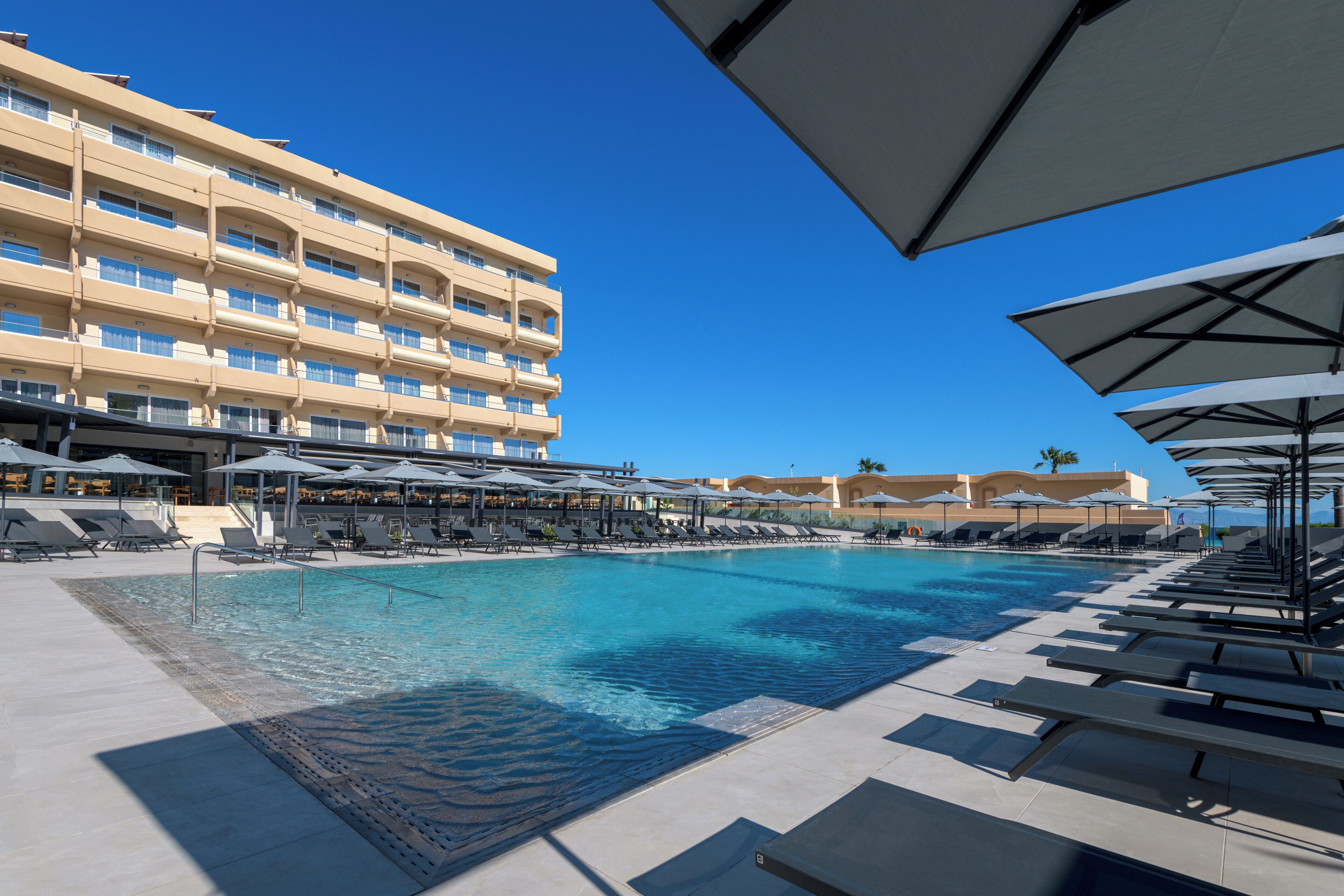 A swimming pool with lounge chairs and umbrellas, a multi-story hotel building in the background, and clear blue skies.