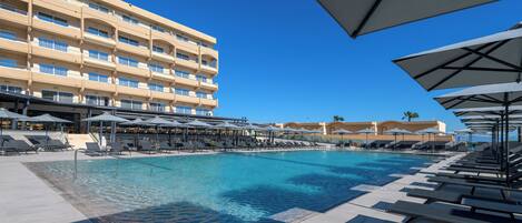 A swimming pool with lounge chairs and umbrellas, a multi-story hotel building in the background, and clear blue skies.