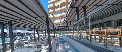 A poolside area with lounge chairs, tables, and umbrellas, overlooking a pool and a building with balconies.