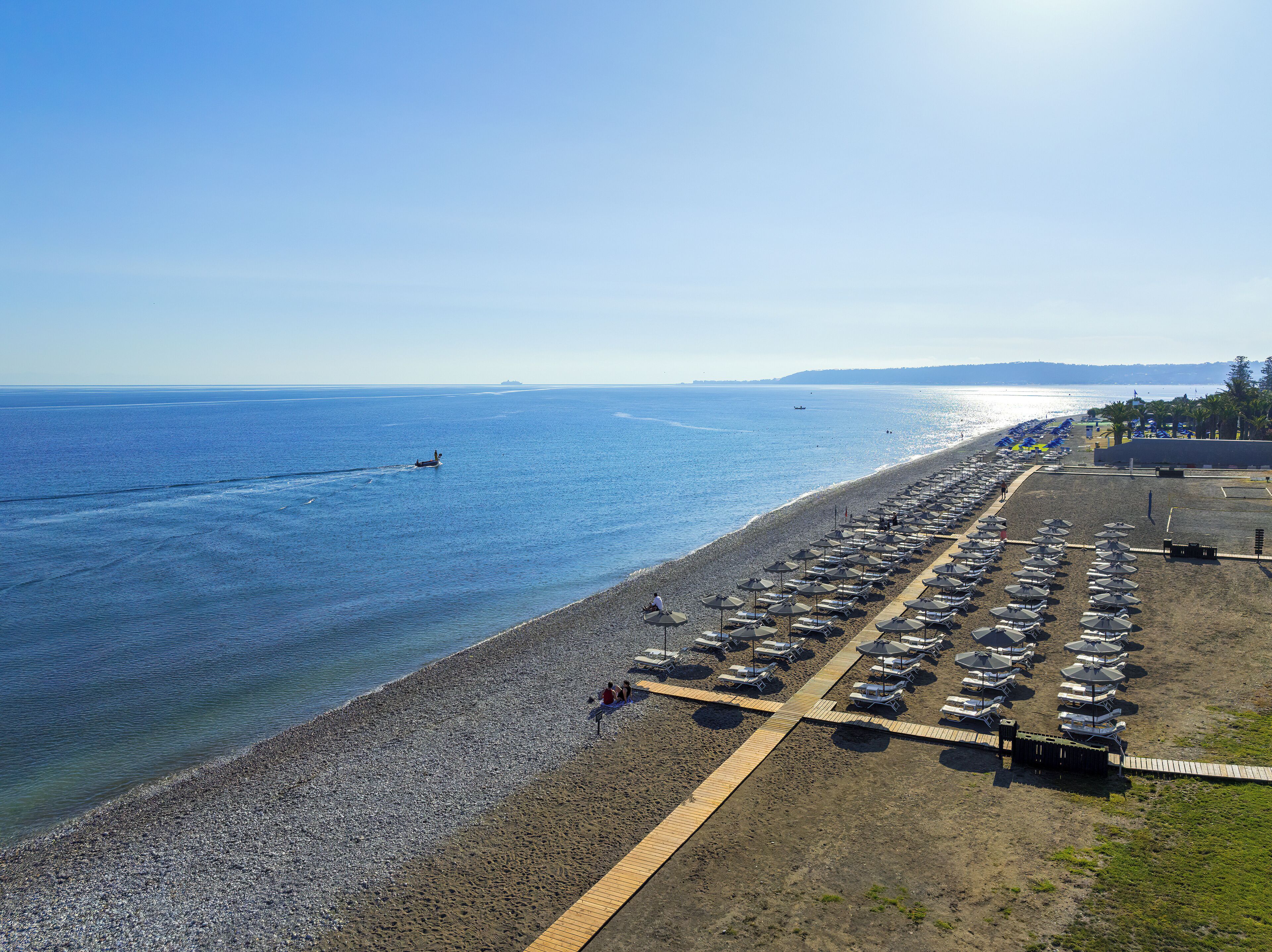 A beach with a row of sunbeds and umbrellas, a wooden walkway, and a boat on the water.