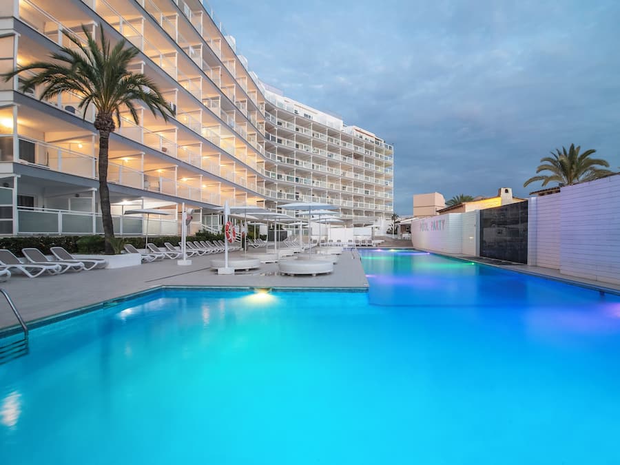 Un hotel con piscina, tumbonas y un edificio de varios pisos con balcones.