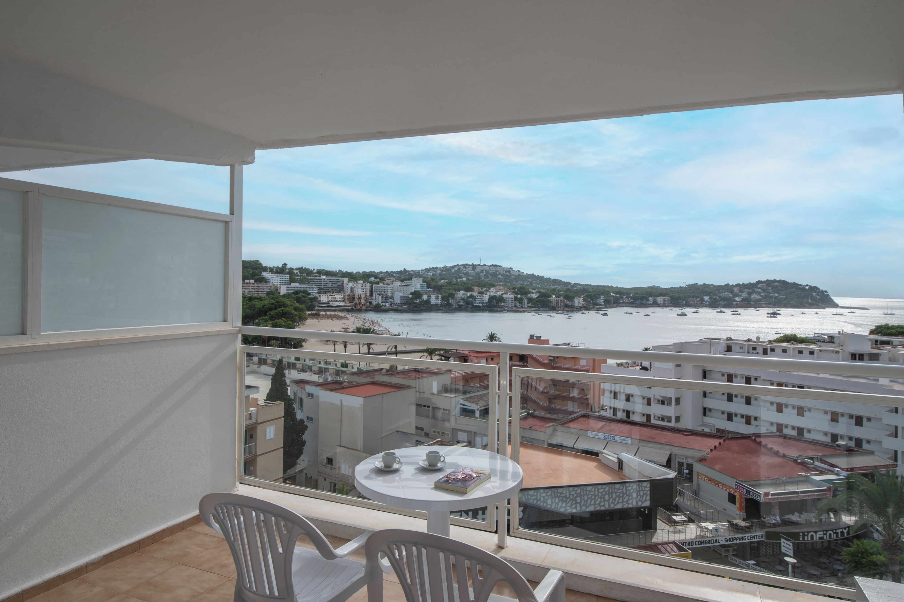 A balcony with a table and chairs, overlooking a beach and a commercial area with buildings and shops.