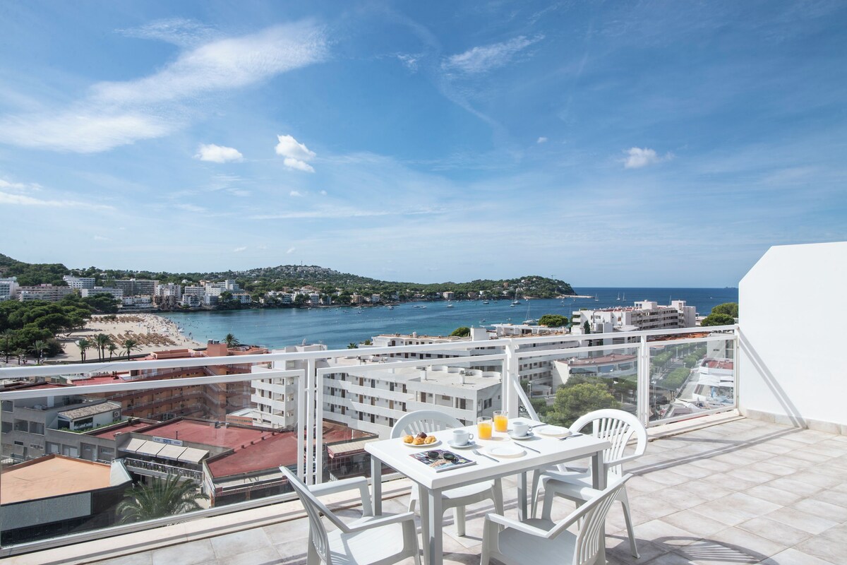 A balcony with a table set for breakfast overlooking a coastal town with buildings and a beach.