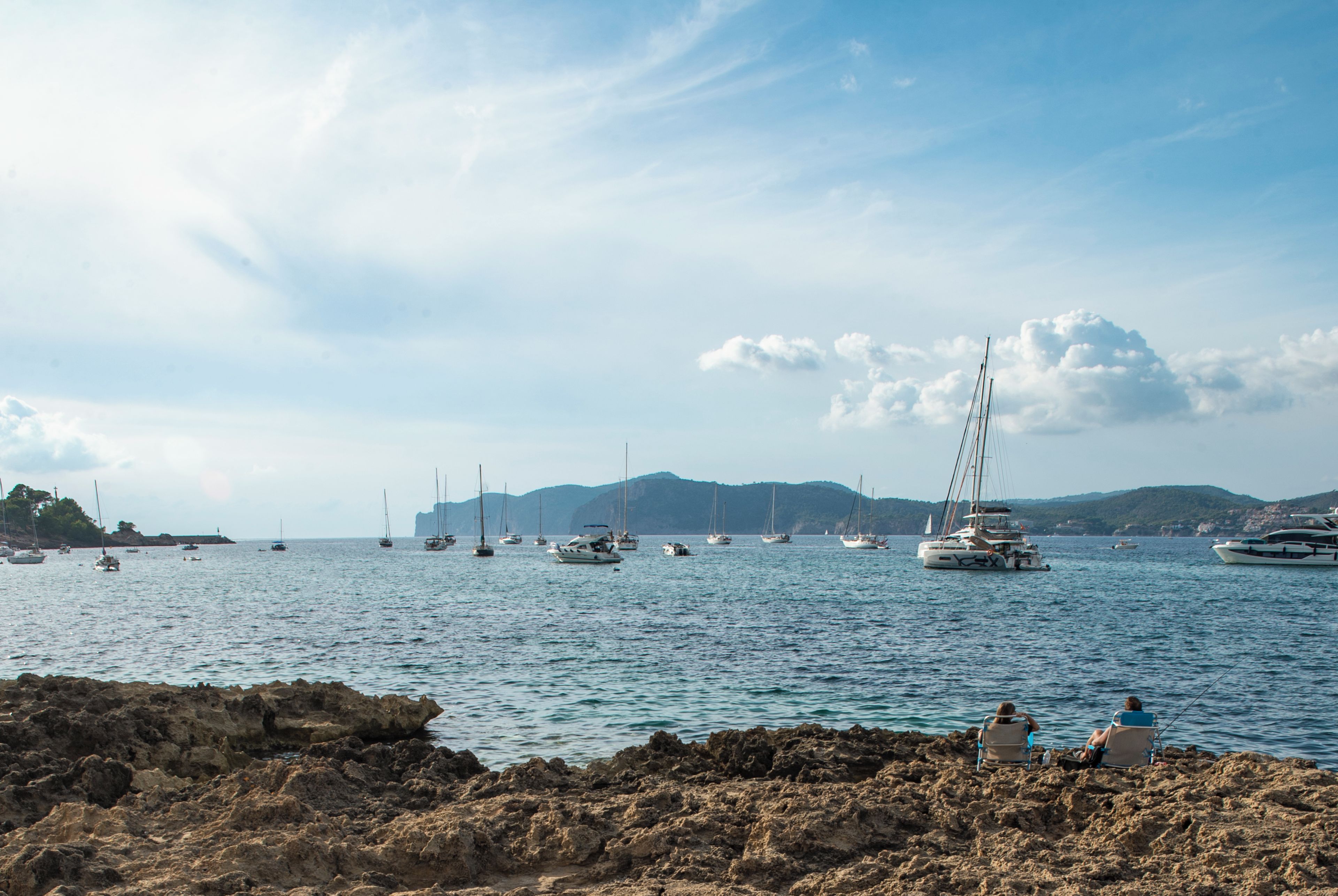 Una costa rocosa con barcos en el agua y colinas al fondo.