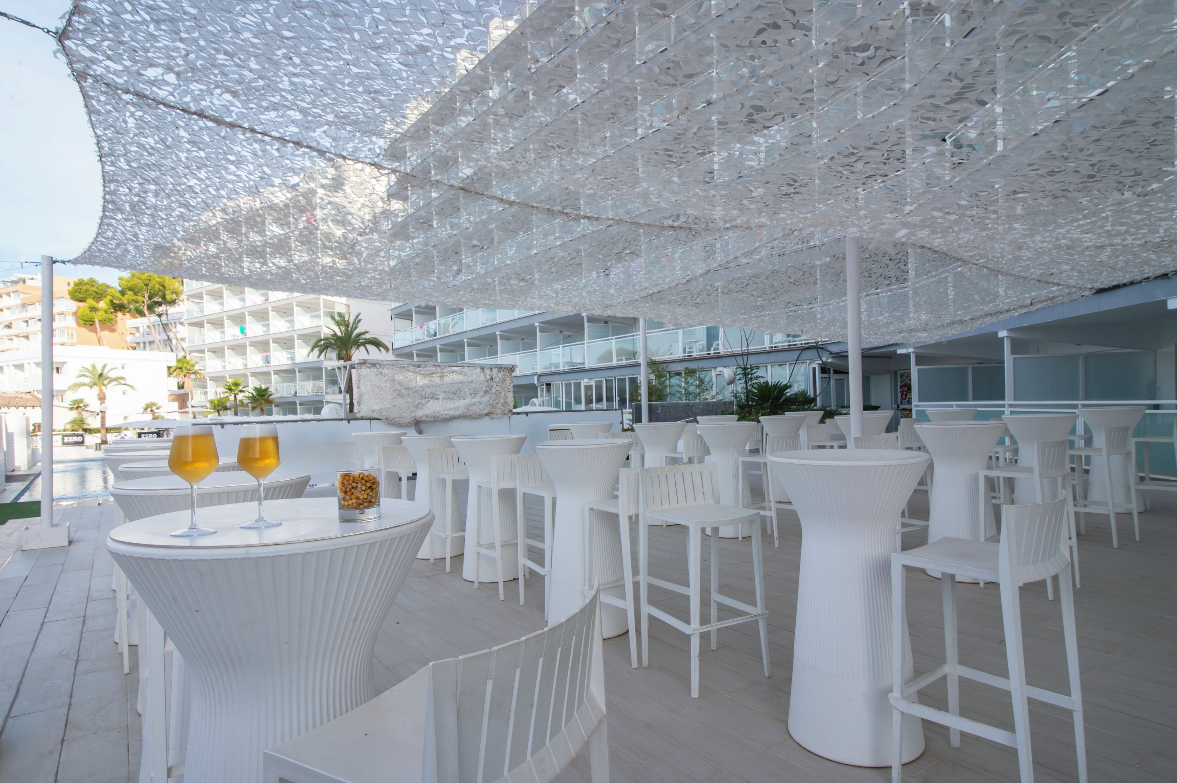 Un espace extérieur aménagé avec des tables rondes et des chaises blanches, une piscine en arrière-plan et une façade de bâtiment moderne.