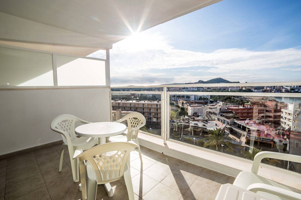 A balcony with a table and chairs, offering a view of a cityscape with buildings and a distant hill.