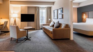 Habitación de hotel con sofá, sillón, mesita, cama, televisión y ventana con cortinas.