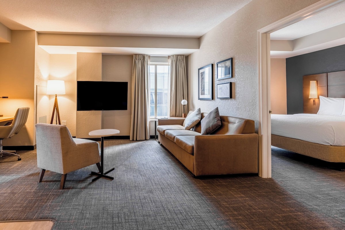 A hotel room with a sofa, a chair, a small table, a bed, a TV, and a window with curtains.
