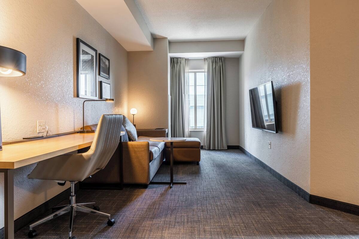 A hotel room with a desk, chair, sofa, TV, and a window with curtains.