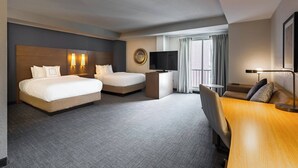 Habitación de hotel con dos camas, un escritorio, un televisor y un ventanal grande con cortinas.