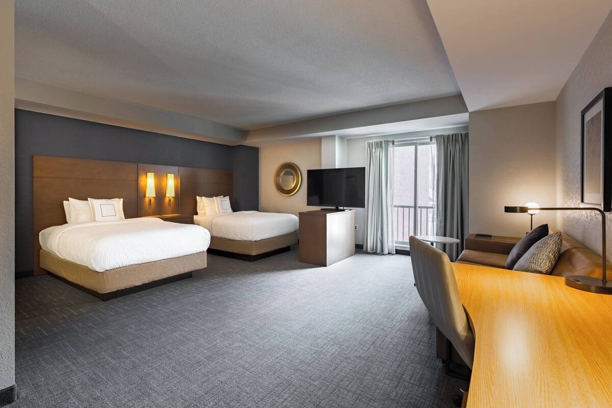 A hotel room with two beds, a desk, a TV, and a large window with curtains.